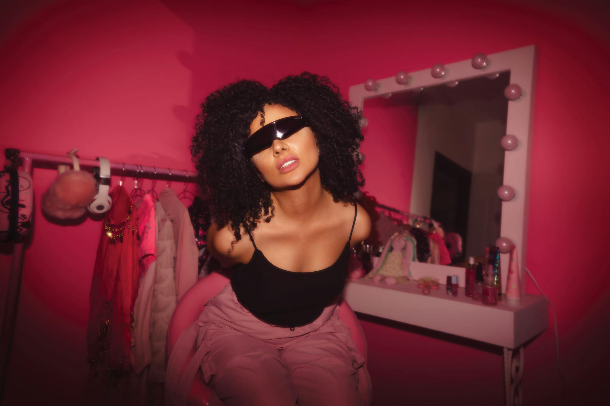 Woman with curly hair and sunglasses posing stylishly in a vibrant pink room.